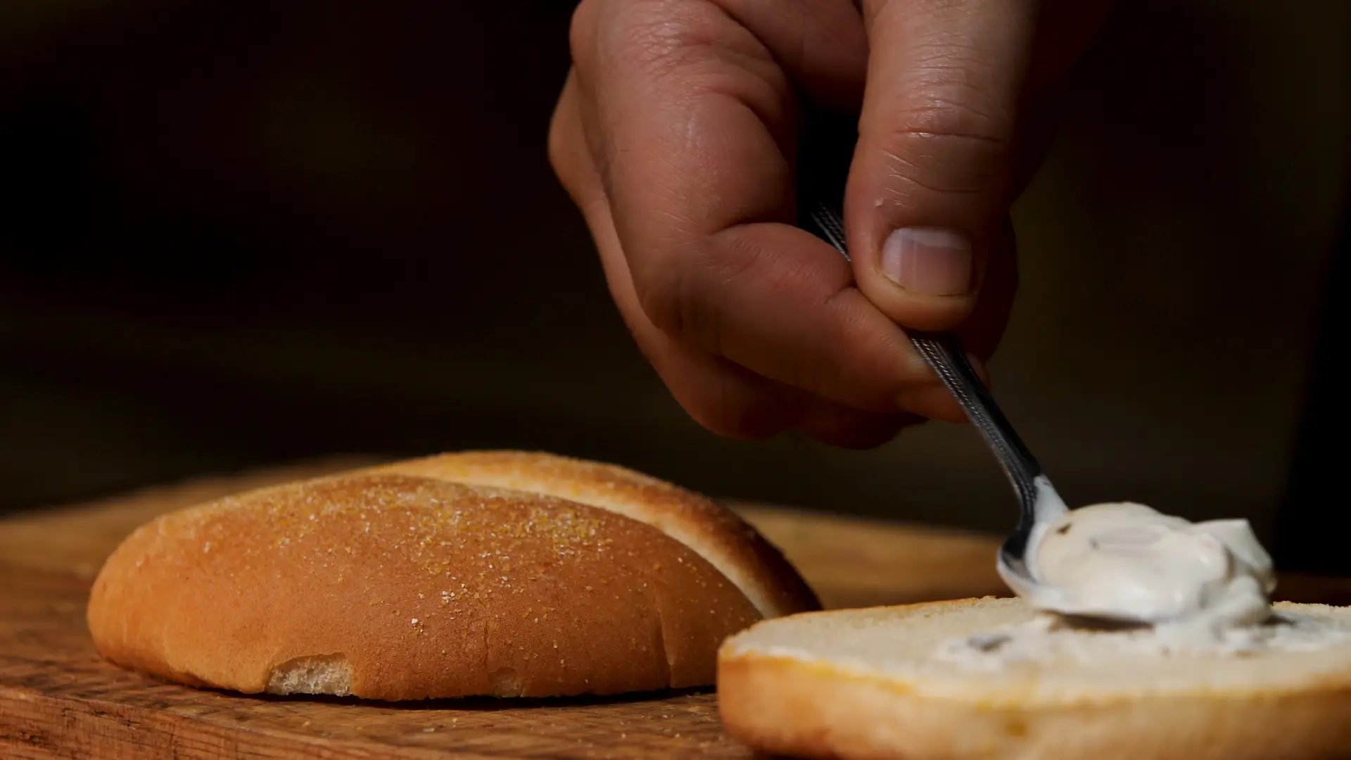 Close up of a person's hand using a spoon to spread white creamy sauce onto a toasted burger bun on a wooden cutting board.