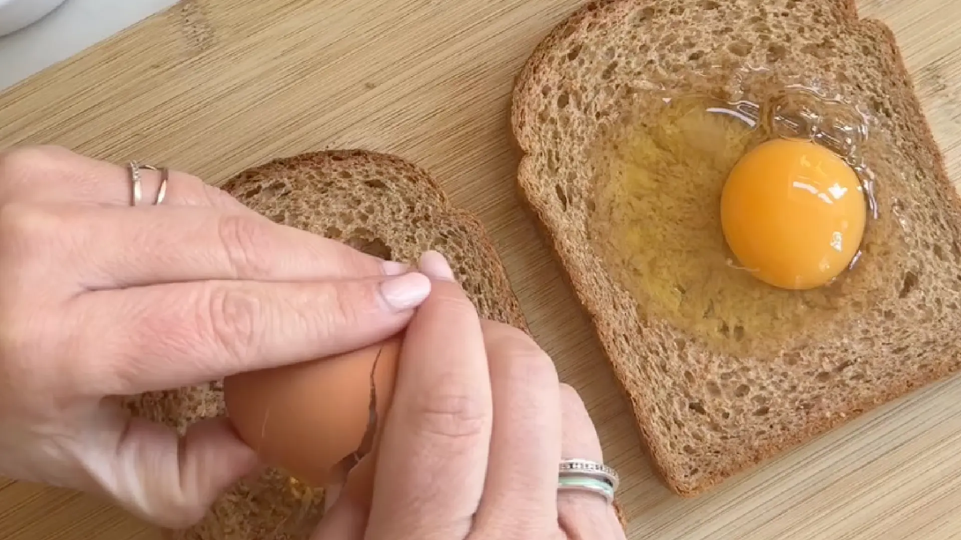 Cracking a fresh egg into the centre indent of a wholemeal bread slice, with another egg already sitting in the adjacent slice.