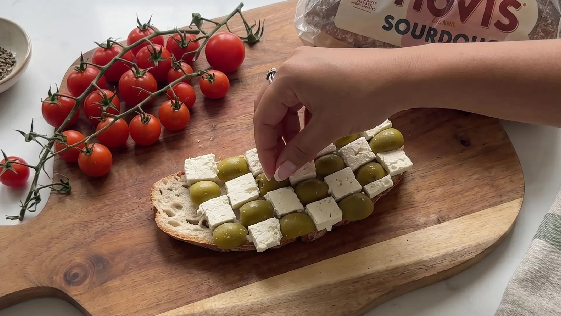 A hand placing a green olive onto a checkerboard pattern of feta cubes and olives on a slice of sourdough toast.