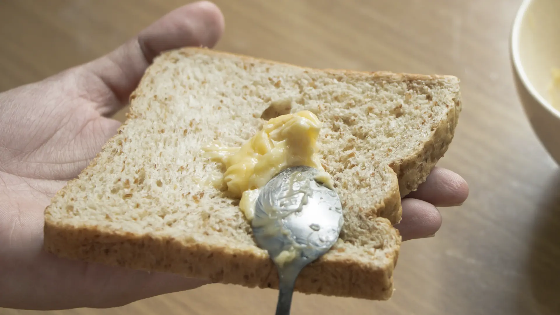 Close-up of a hand holding a slice of wholemeal granary bread while spreading soft butter onto it with a spoon.
