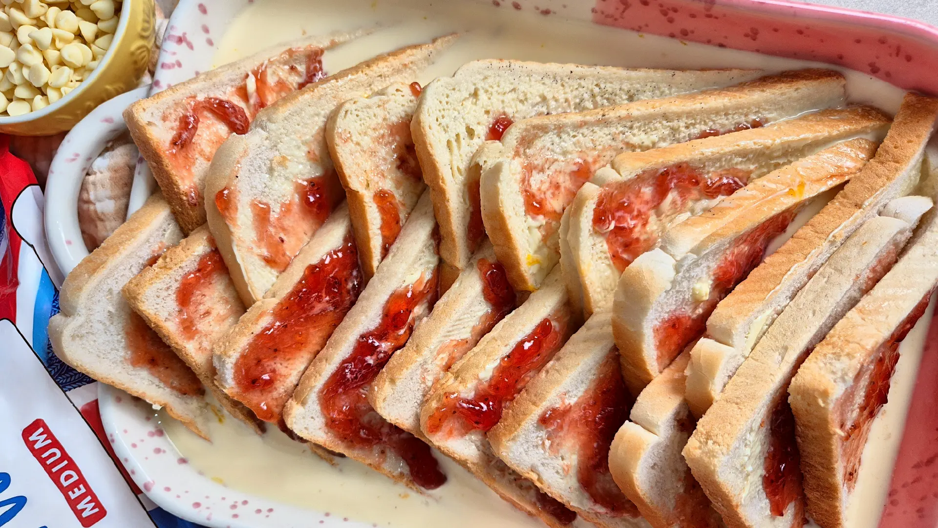 Triangular slices of jam spread bread soaking in a baking dish filled with a light custard mixture.