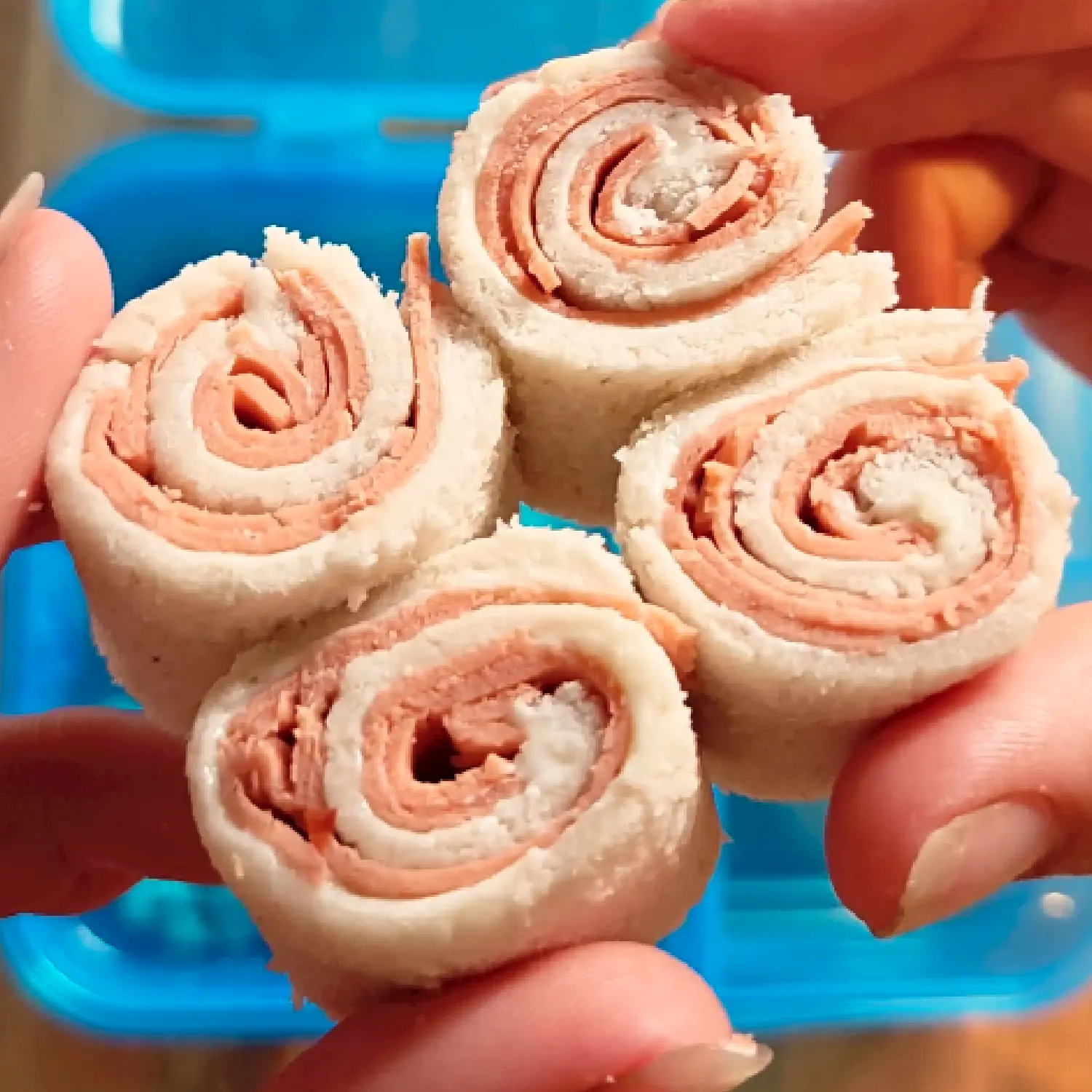 Hands holding four rolled ham and cream cheese pinwheel sandwich bite size pieces over a blue lunchbox.