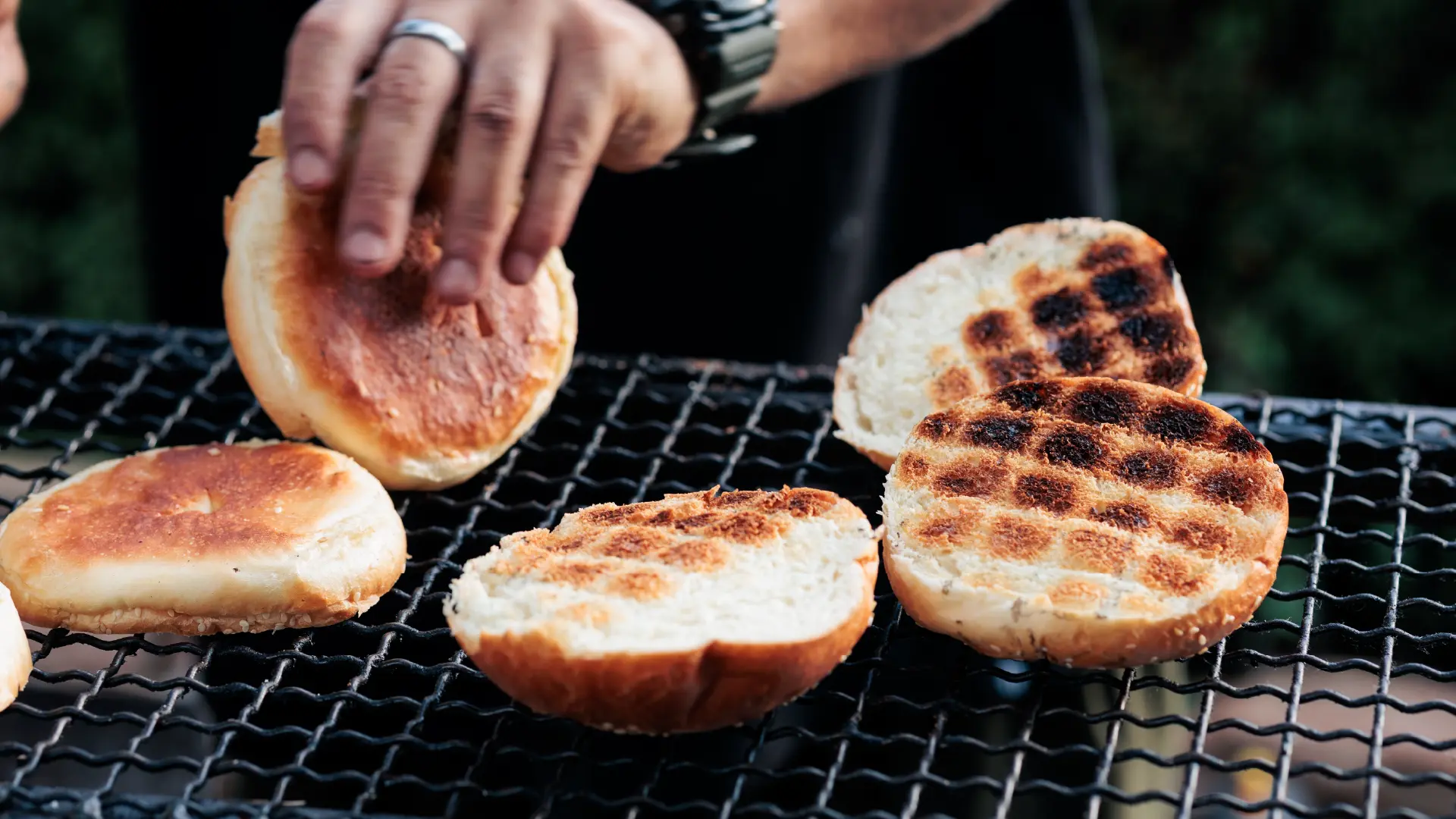 A hand reaches down to touch hamburger buns toasting with grid char marks on an outdoor barbecue grill.