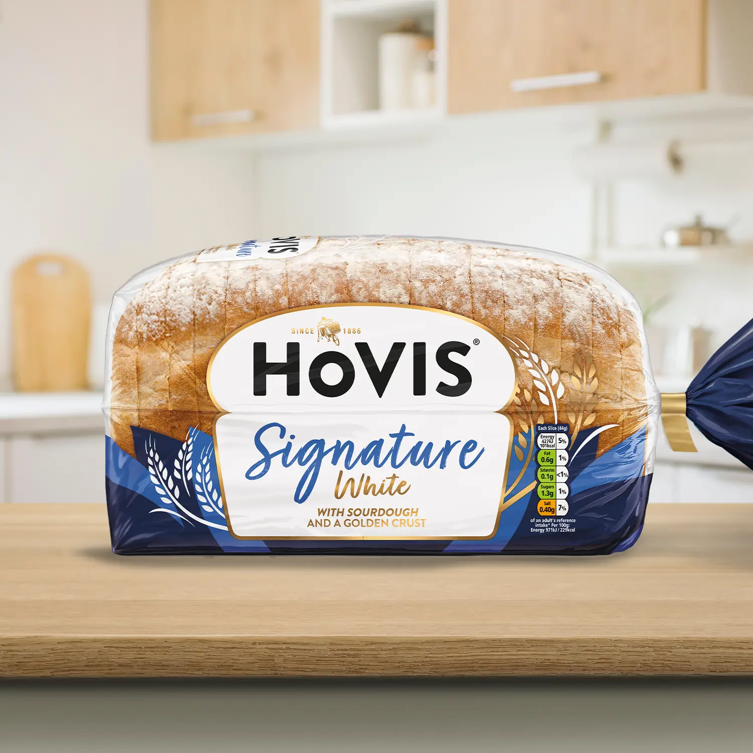 A loaf of Hovis Signature White bread with sourdough and a crust dusted with flour, displayed on a wooden kitchen countertop. The packaging features a white and navy design with a gold trim and nutrition information. 