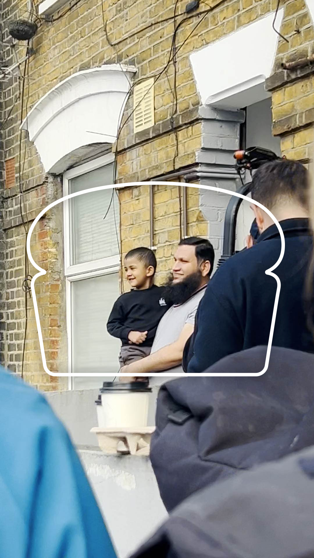 
A vertical shot framed by a white outline resembling a slice of bread. Inside the frame, a man with a long beard smiles while holding a young child, who is also smiling and looking off to the left. They are positioned in front of a window on a brick building, with another person partially visible to the right. The foreground is blurred with the shoulders of someone in a blue jacket and dark clothing. The brick building and a white archway above the window are visible in the background.