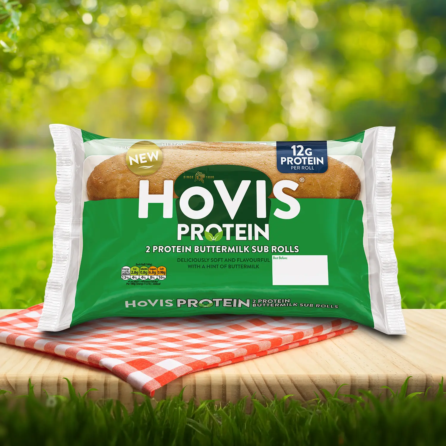 Hovis Protein Buttermilk Sub Rolls packaging on a picnic table with a green outdoor background. The pack shows two soft sub rolls with 12g of protein per roll and highlights “Deliciously soft and flavourful with a hint of buttermilk.” A red gingham cloth adds a picnic feel to the setting.
