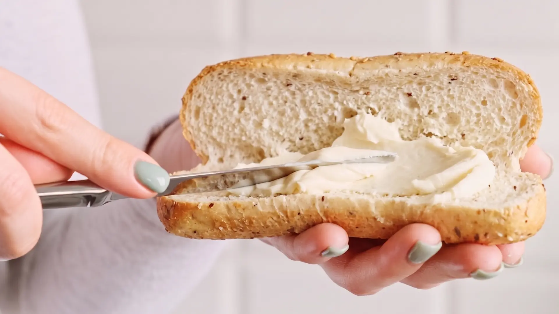 A person uses a butter knife to spread creamy white cheese onto a sliced multi-seed baguette roll.