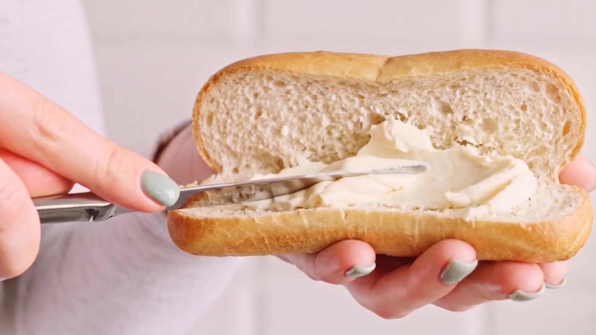 A person uses a butter knife to spread creamy white cheese onto a sliced baguette roll.