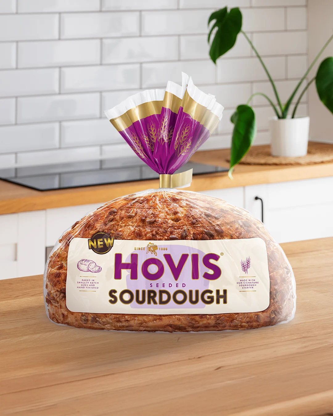 A loaf of Hovis Seeded Sourdough bread displayed on a wooden kitchen countertop. The packaging features a white and purple design with a gold trim and nutrition information. A 'NEW' label is shown on the top corner of the pack