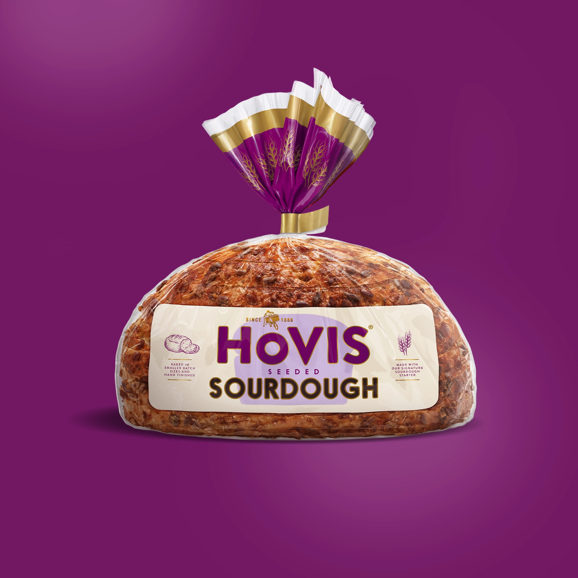 A loaf of Hovis Seeded Sourdough bread is displayed on a purple background. The packaging features a white and purple design with a gold trim and nutrition information.
