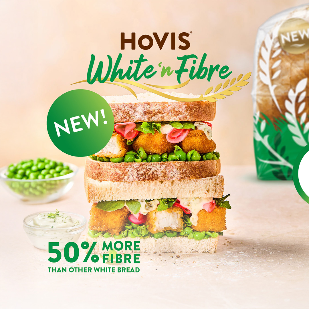 A layered sandwich made with Hovis White 'n' Fibre bread takes centre stage. The sandwich is filled with golden-brown fish fingers, green peas, pink pickled onions, and a creamy white sauce, with some green leaves peeking out. To the left, a small bowl of green peas and a dish of white sauce are visible. In the bottom left corner, large green text reads "50% MORE FIBRE" with smaller text below saying "THAN OTHER WHITE BREAD". A bright green circle with "NEW!" in white text overlaps the top left of the sandwich. At the top, the Hovis logo is present, with "White 'n' Fibre" written in a flowing green script above images of wheat. The background is a soft, light brown, suggesting a natural or wholesome feel.