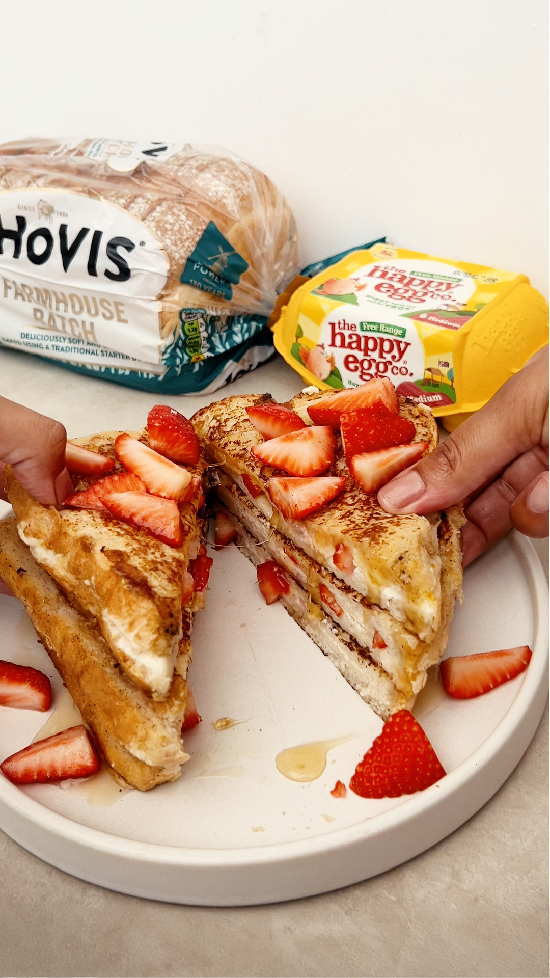 A plate of French toast made with Hovis Farmhouse Batch bread, layered with cream and sliced strawberries, and topped with more fresh strawberries and a drizzle of syrup. Two hands hold each half of the sandwich. In the background, a loaf of Hovis Farmhouse Batch bread and a carton of The Happy Egg Co. free-range eggs are visible