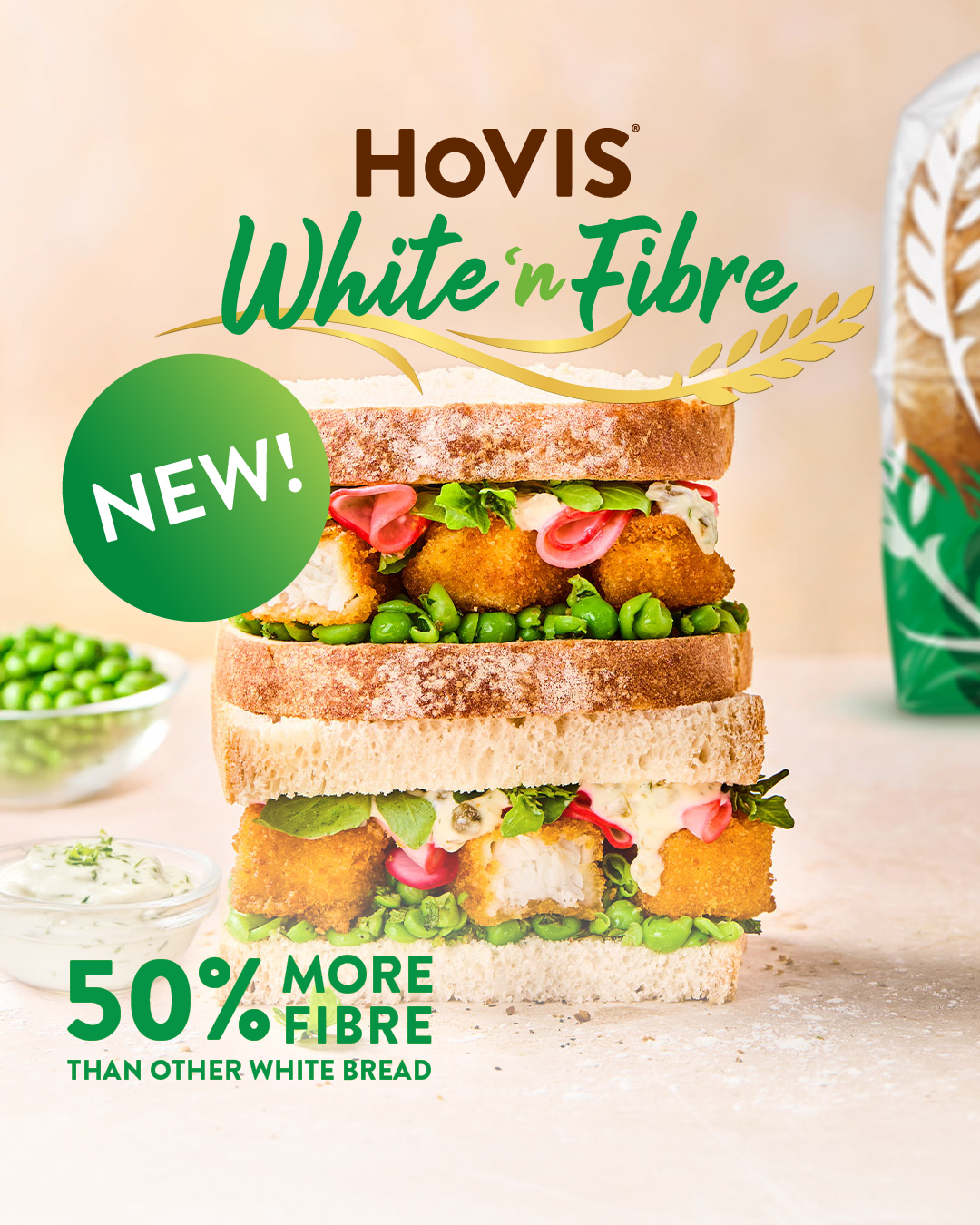 
A layered sandwich made with Hovis White 'n' Fibre bread takes centre stage. The sandwich is filled with golden-brown fish fingers, green peas, pink pickled onions, and a creamy white sauce, with some green leaves peeking out. To the left, a small bowl of green peas and a dish of white sauce are visible. In the bottom left corner, large green text reads "50% MORE FIBRE" with smaller text below saying "THAN OTHER WHITE BREAD". A bright green circle with "NEW!" in white text overlaps the top left of the sandwich. At the top, the Hovis logo is present, with "White 'n' Fibre" written in a flowing green script above images of wheat. The background is a soft, light brown, suggesting a natural or wholesome feel.
