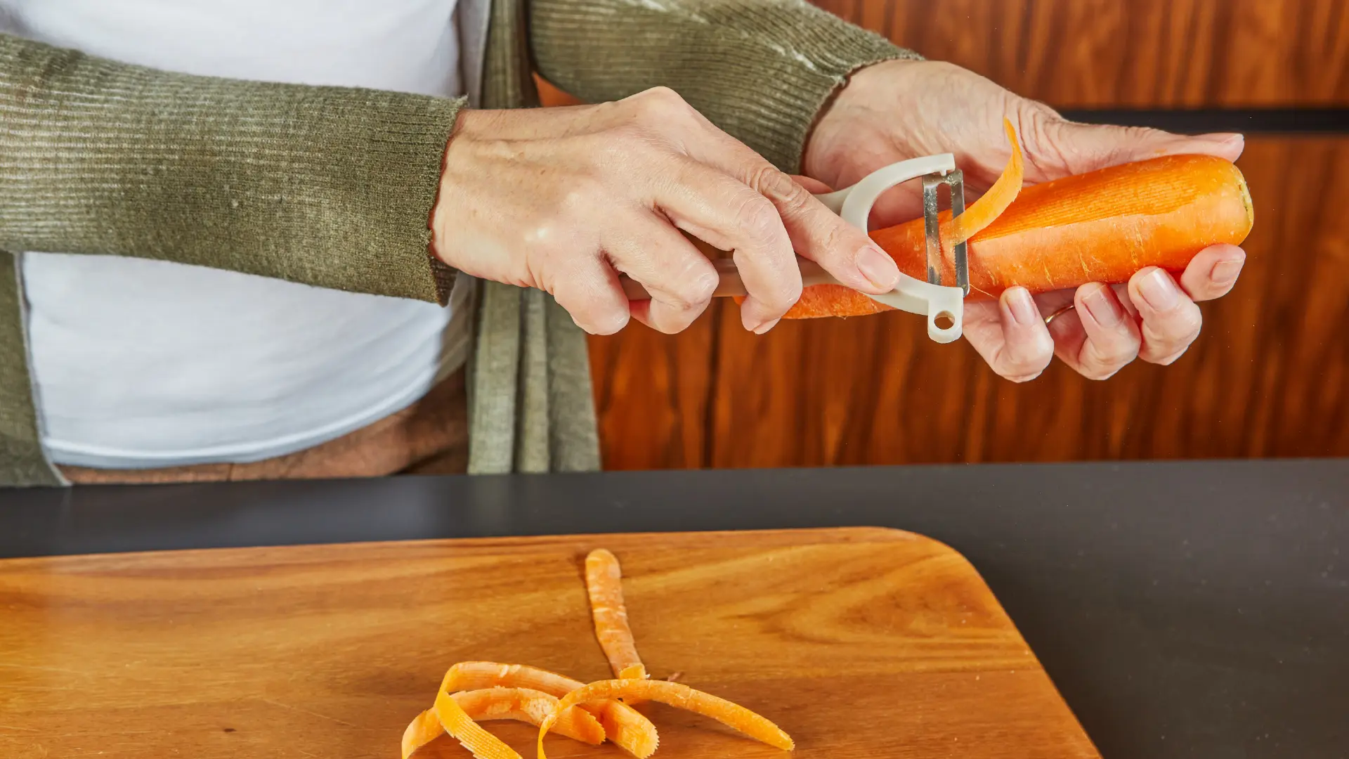 A person uses a Y-shaped peeler to remove the skin from a bright orange carrot over a wooden cutting board.