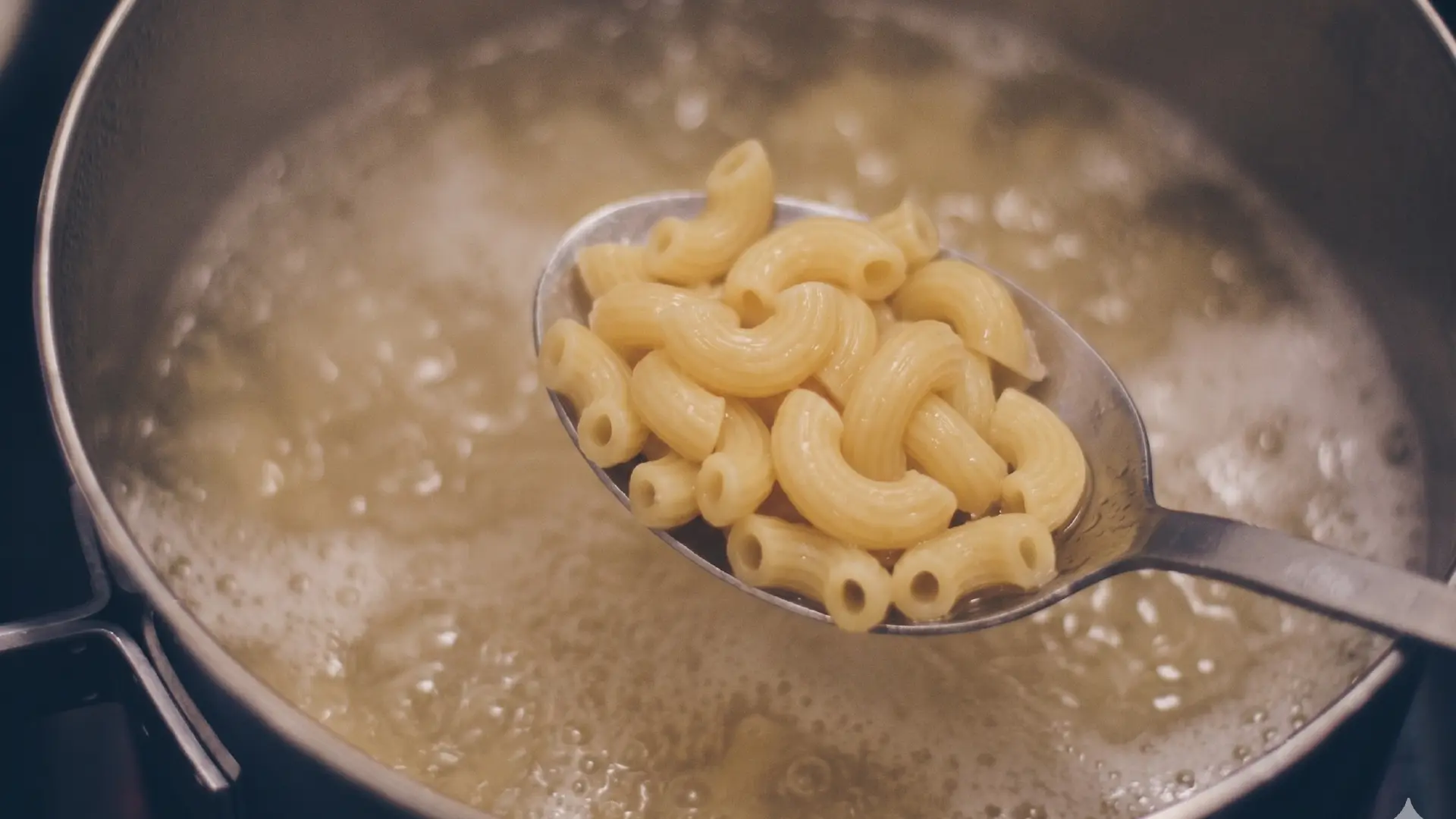 A metal spoon lifts cooked macaroni from a pot of boiling water, with steam rising from the bubbling surface.