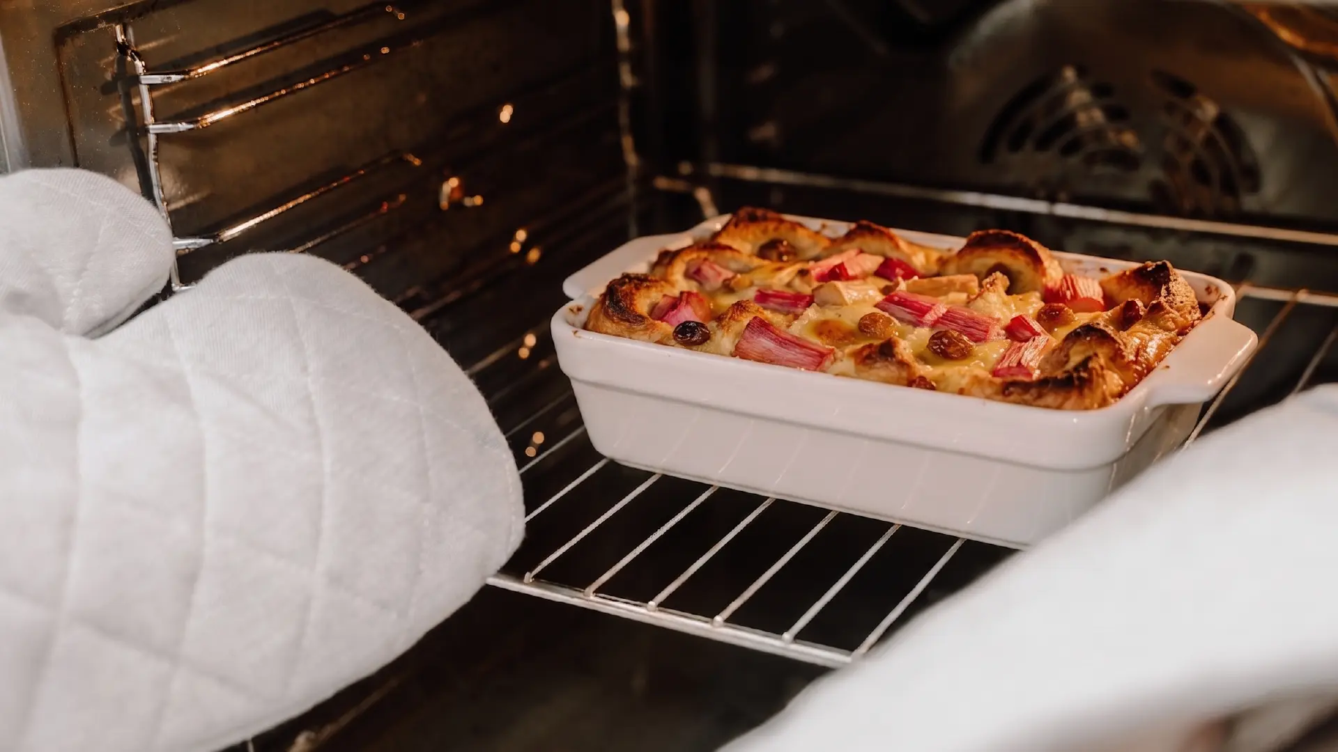 A white dish of golden-brown rhubarb bread pudding sits on an oven rack, framed by two white quilted oven mitts.