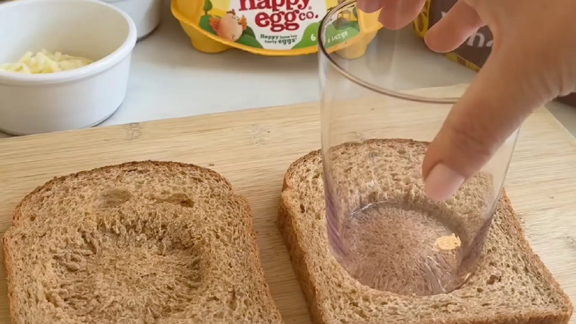 Using a glass to press a circular indent into thick wholemeal bread slices on a wooden cutting board for an egg recipe.