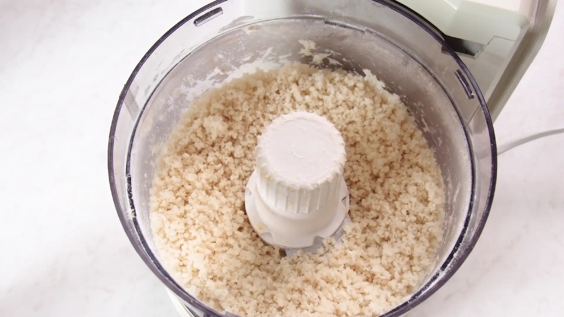 Top-down view of a clear food processor bowl filled with coarsely blended crumbly white breadcrumb mixture.