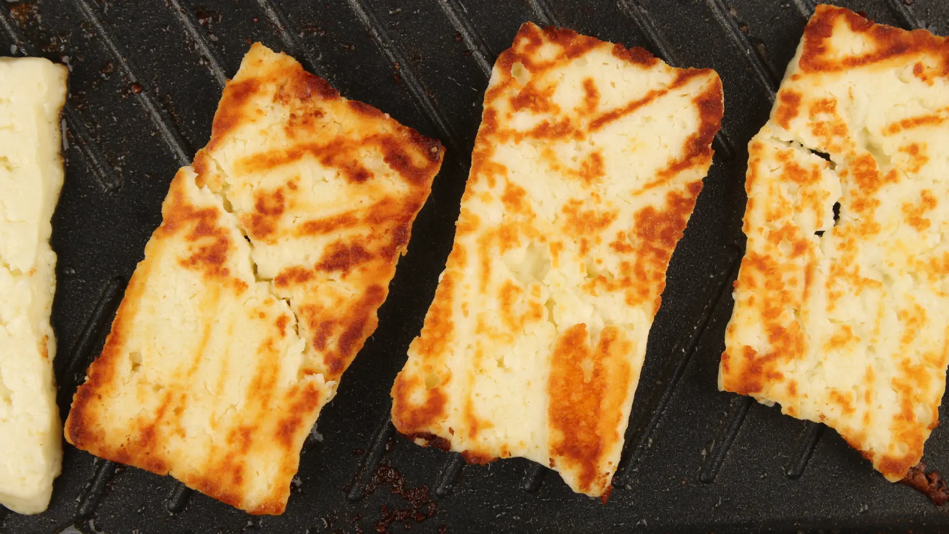 Top down view of rectangular slices of halloumi cheese with golden brown grill marks cooking on a black ridged grill pan.