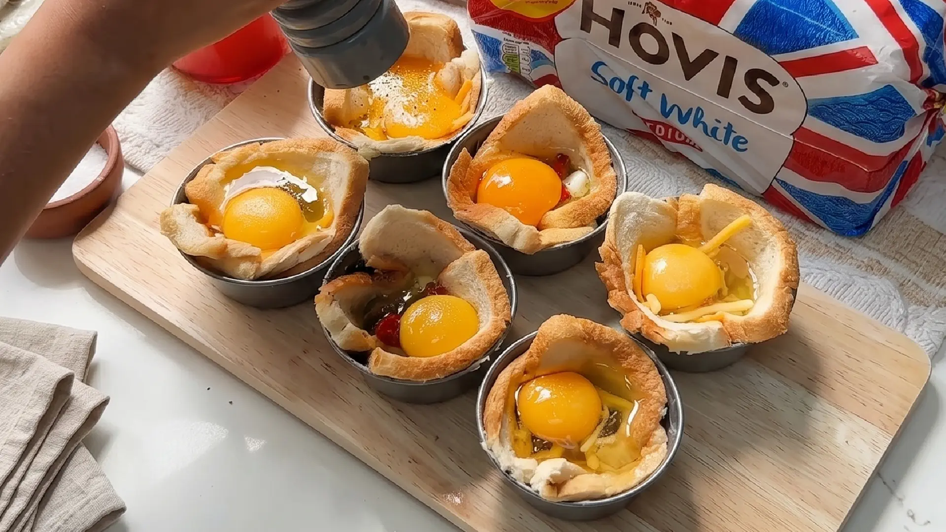 A hand cracks fresh eggs into bread-lined tins, seasoning the yolks for baked breakfast cups on a wooden cutting board.