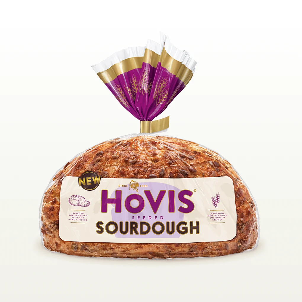 Hovis Seeded Sourdough bread on a white backdrop with a subtle shadow. The packaging is half transparent and half light and dark purple with gold detailing