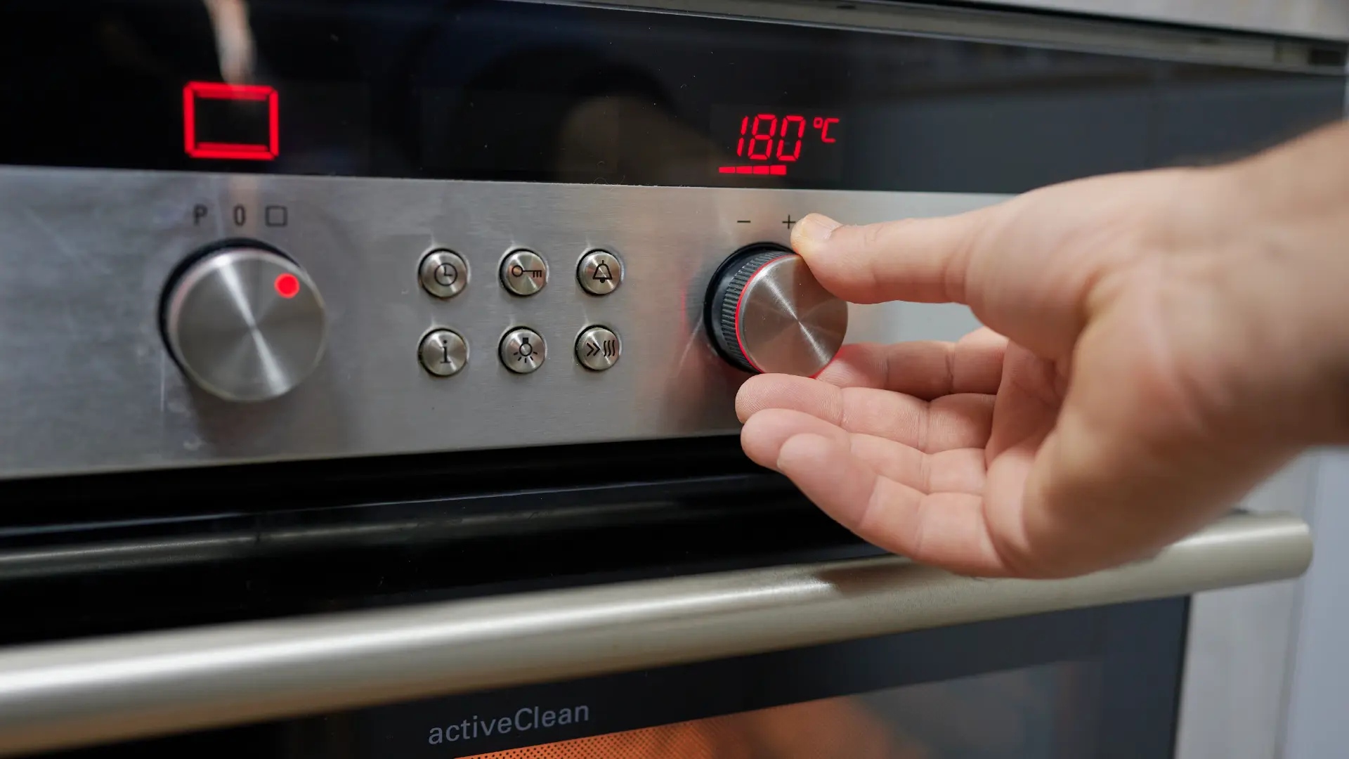 A hand turns the silver knob on a stainless steel oven control panel to set the red digital temperature display to 180°C.