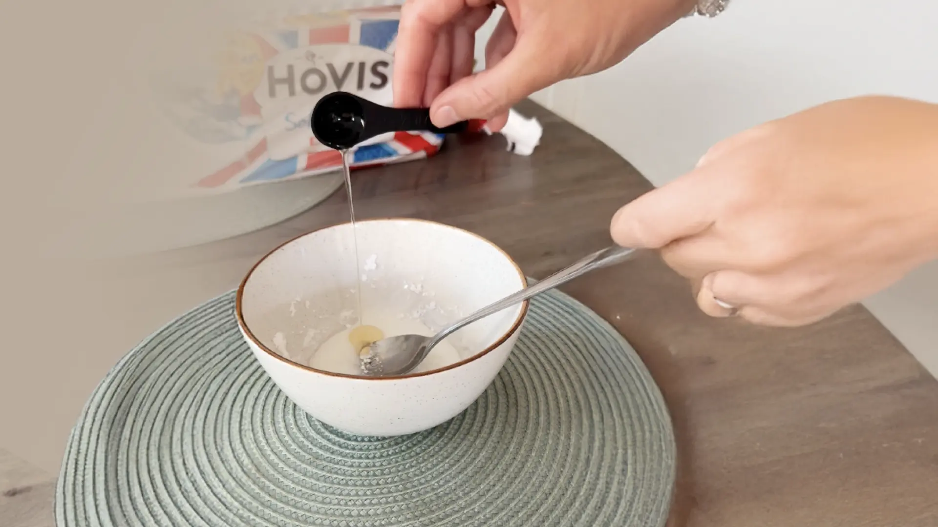 A hand measuring liquid into a white bowl using a black spoon, with a metal spoon inside and Hovis bread in the back.