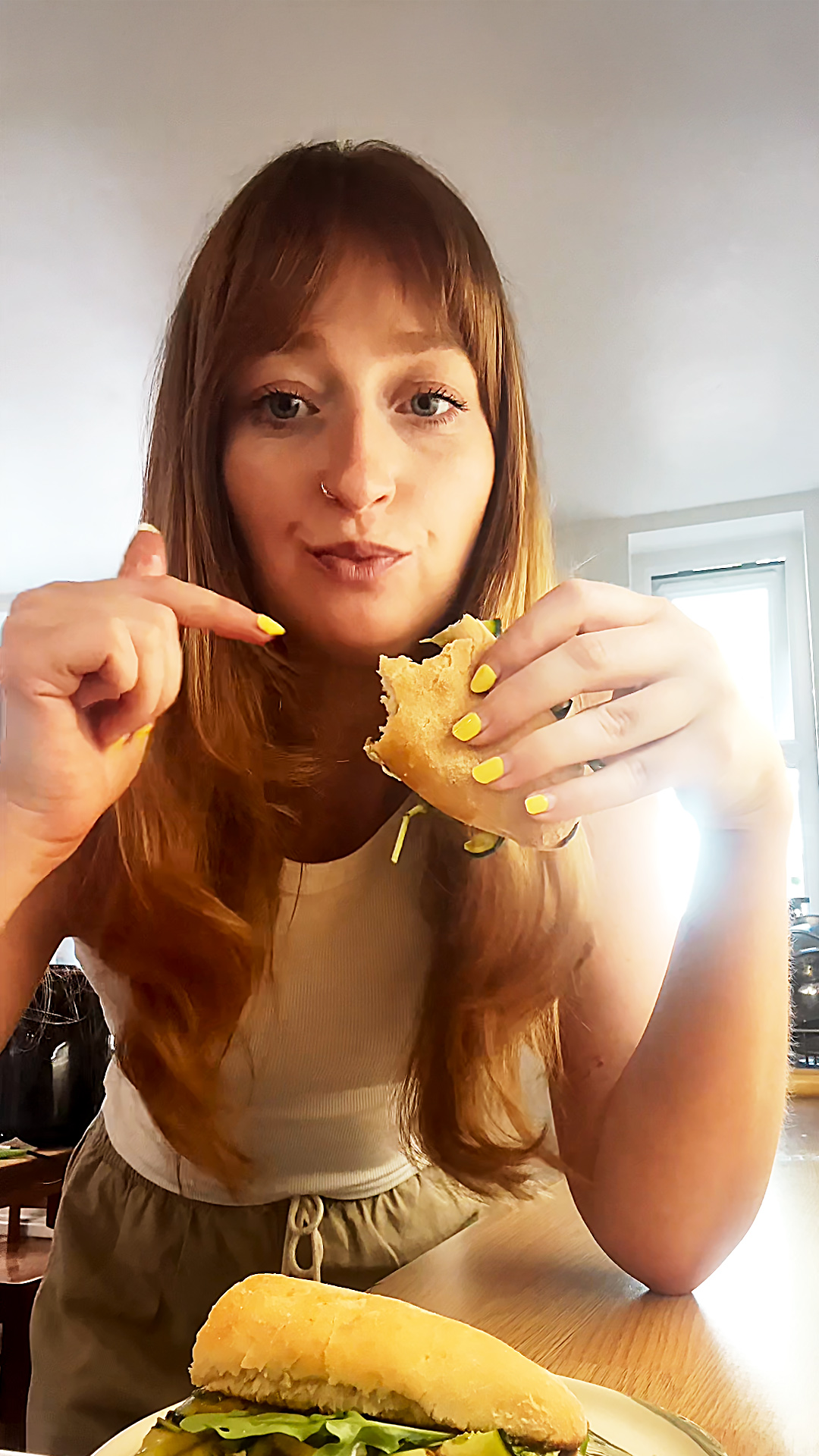 A woman with long reddish-brown hair and yellow-painted nails sits at a table, holding a partially eaten sub and pointing toward it with a smile. She wears a white tank top and beige trousers, with another sandwich on a plate in front of her