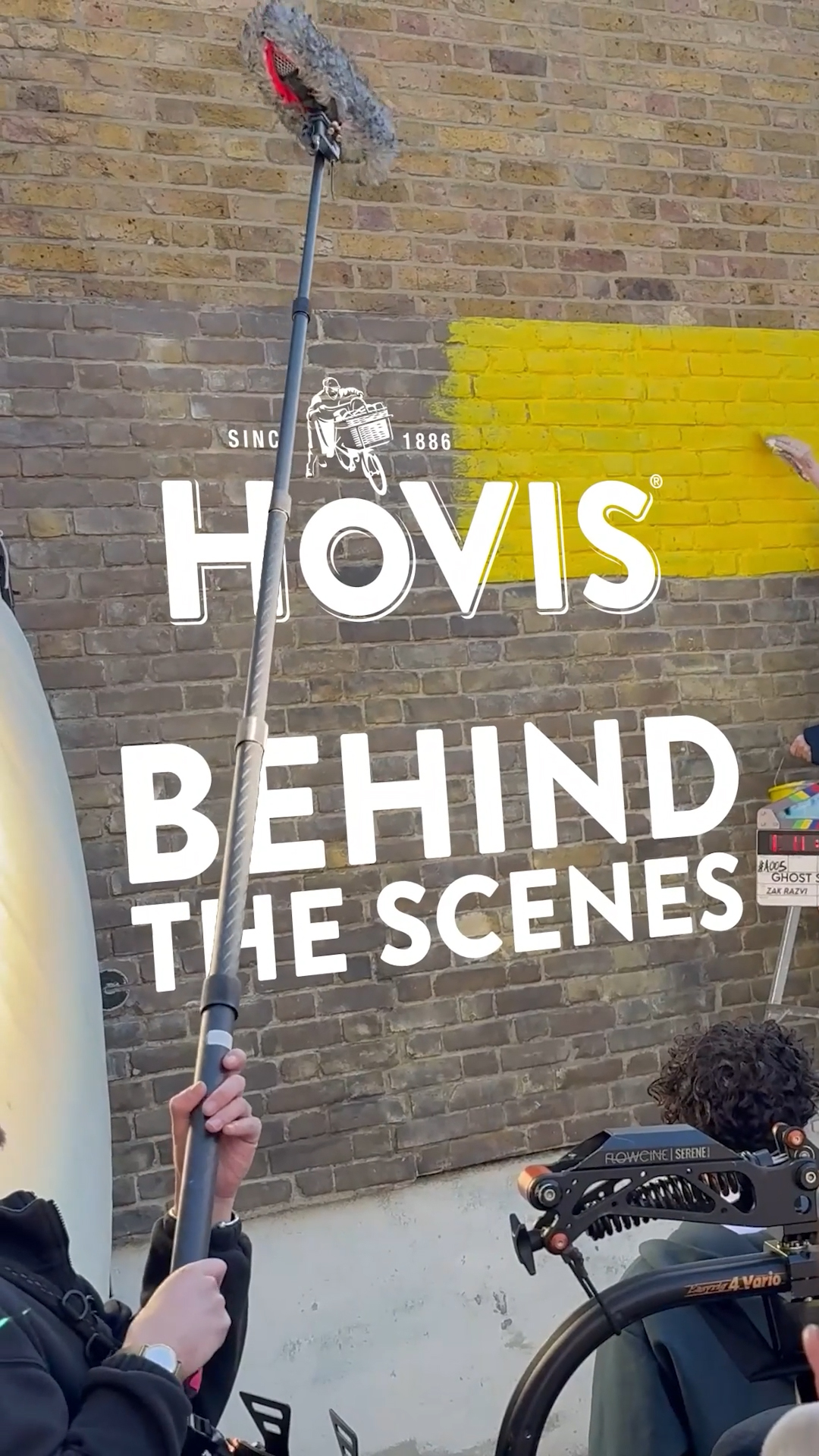 
A behind-the-scenes vertical shot showing a film set against a brick wall. A long boom microphone with a fluffy windsock extends upwards in the centre, held by hands at the bottom. Large white text overlays the image, reading "HOVIS" at the top with a small "SINCE 1886" and a baker on a bicycle logo, followed by "BEHIND THE SCENES" below. The brick wall behind has a bold yellow stripe painted across it. Part of a camera rig with a black curly-haired person behind it is visible in the bottom right corner.
