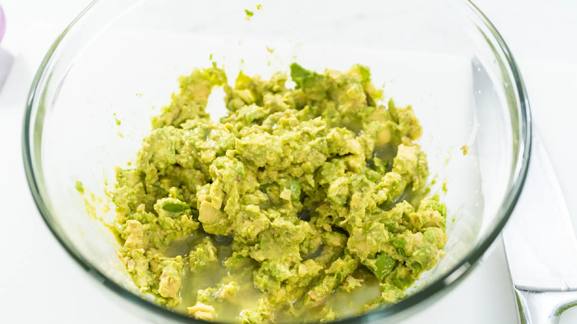 Freshly mashed avocado in a clear glass mixing bowl set against a clean white background.