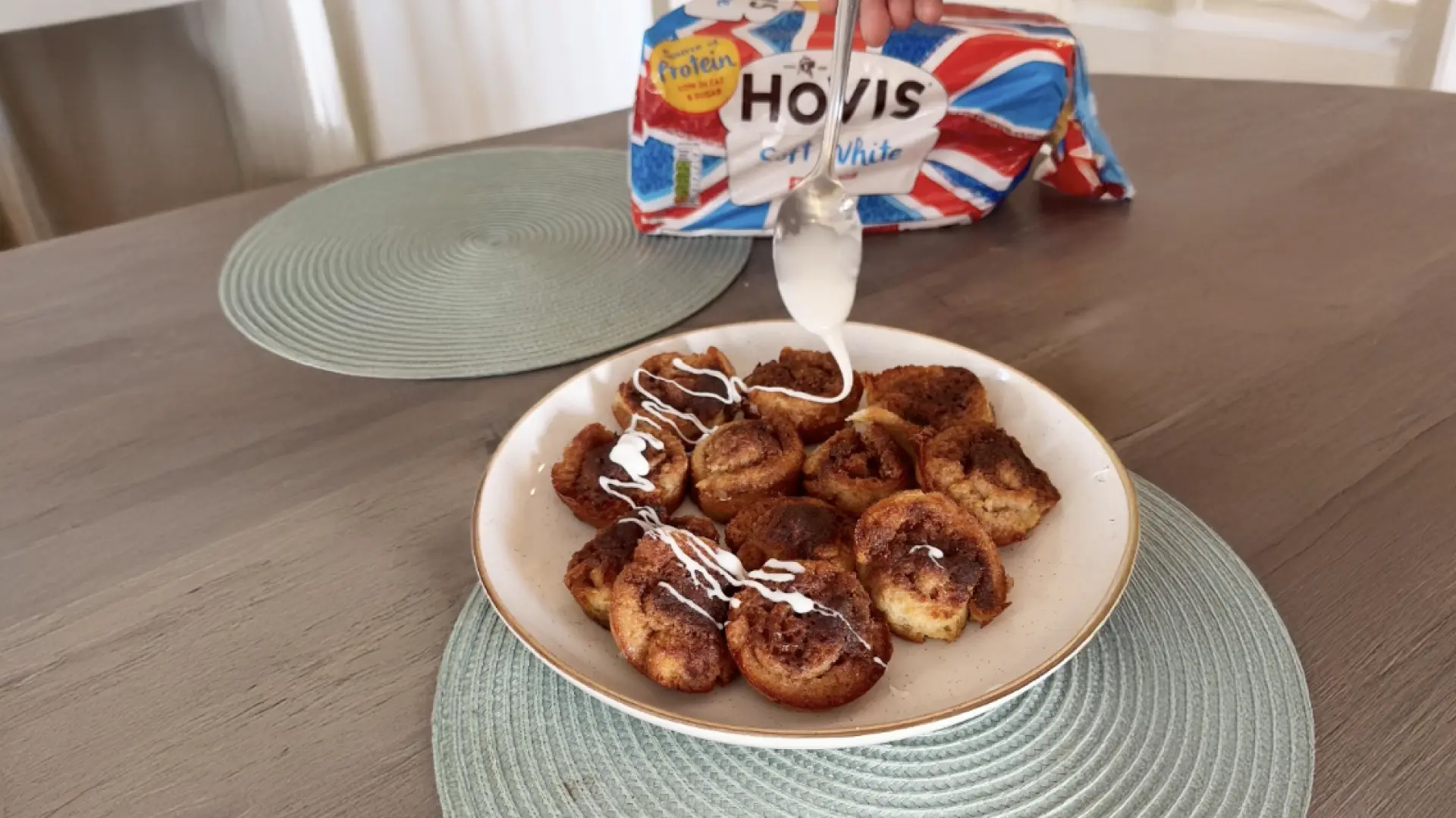 A hand drizzling white icing from a spoon over baked cinnamon rolls on a plate, with Hovis bread in the background.