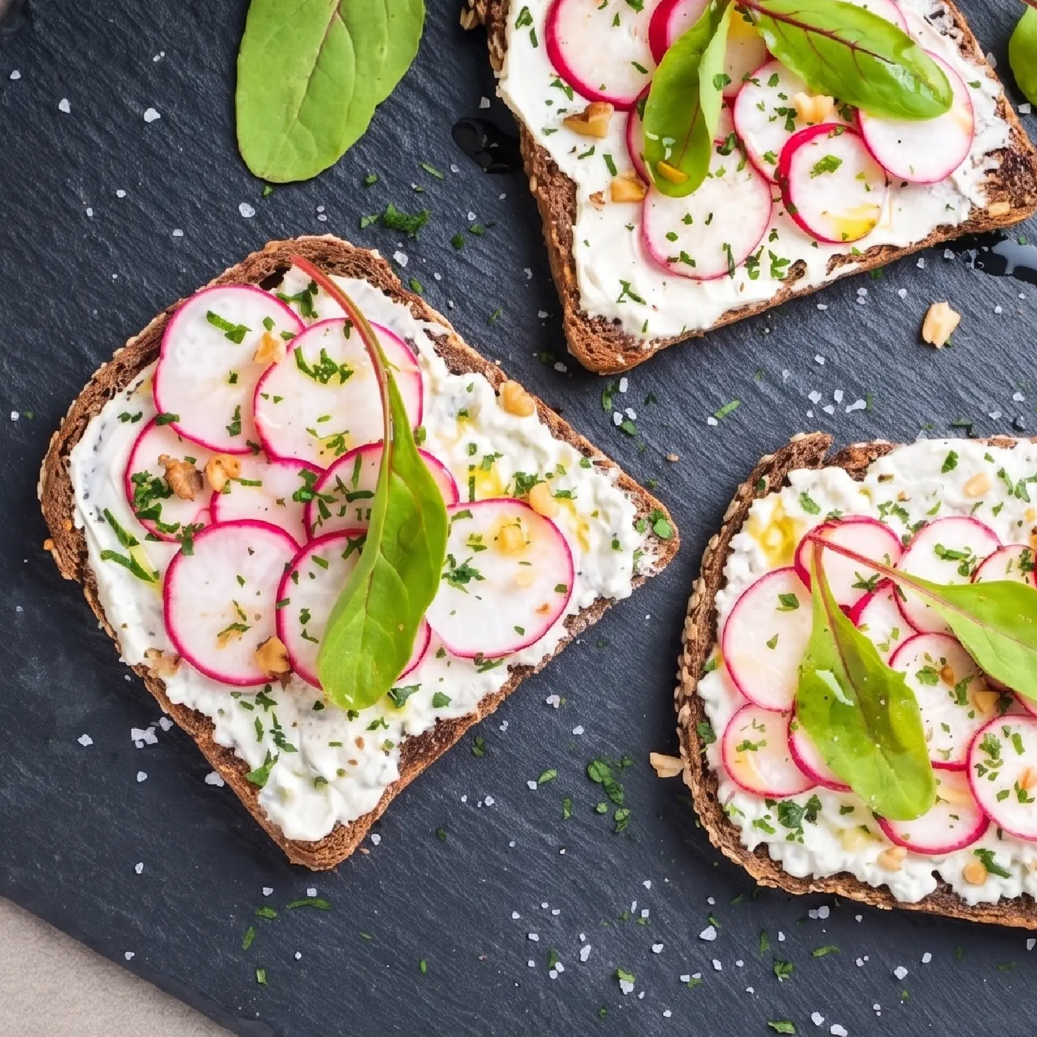 Top-down view of three radish toasts with ricotta, herbs, and greens on a dark slate board with scattered sea salt.