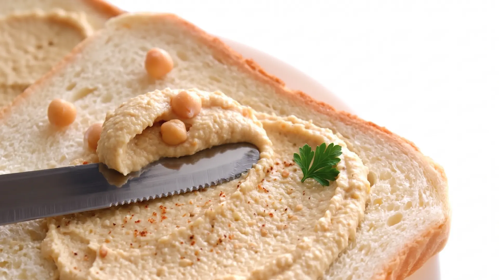 Close up of a butter knife spreading smooth hummus onto a slice of white bread, topped with whole chickpeas and paprika.