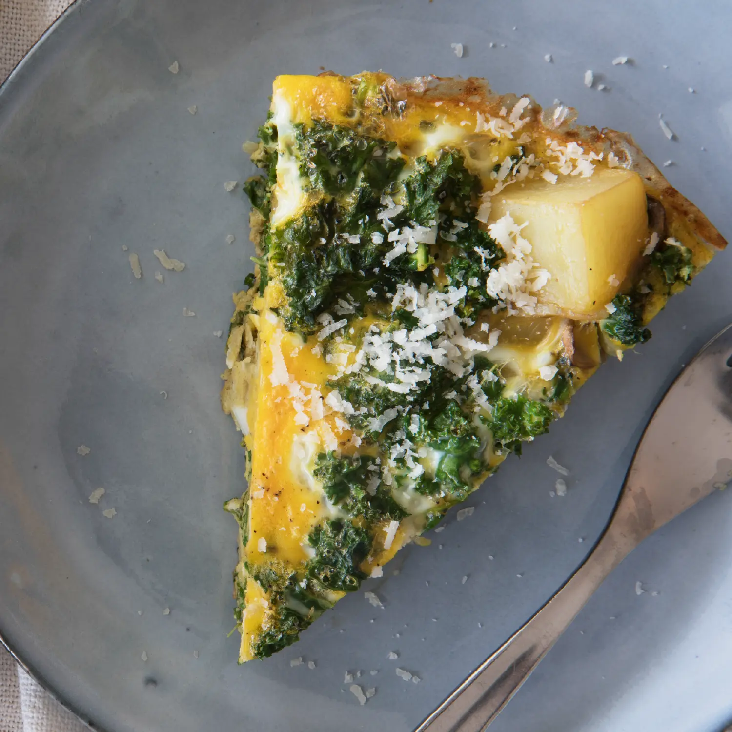 A slice of golden egg frittata with cubed potatoes, topped with green pesto and grated parmesan cheese on a round grey plate.