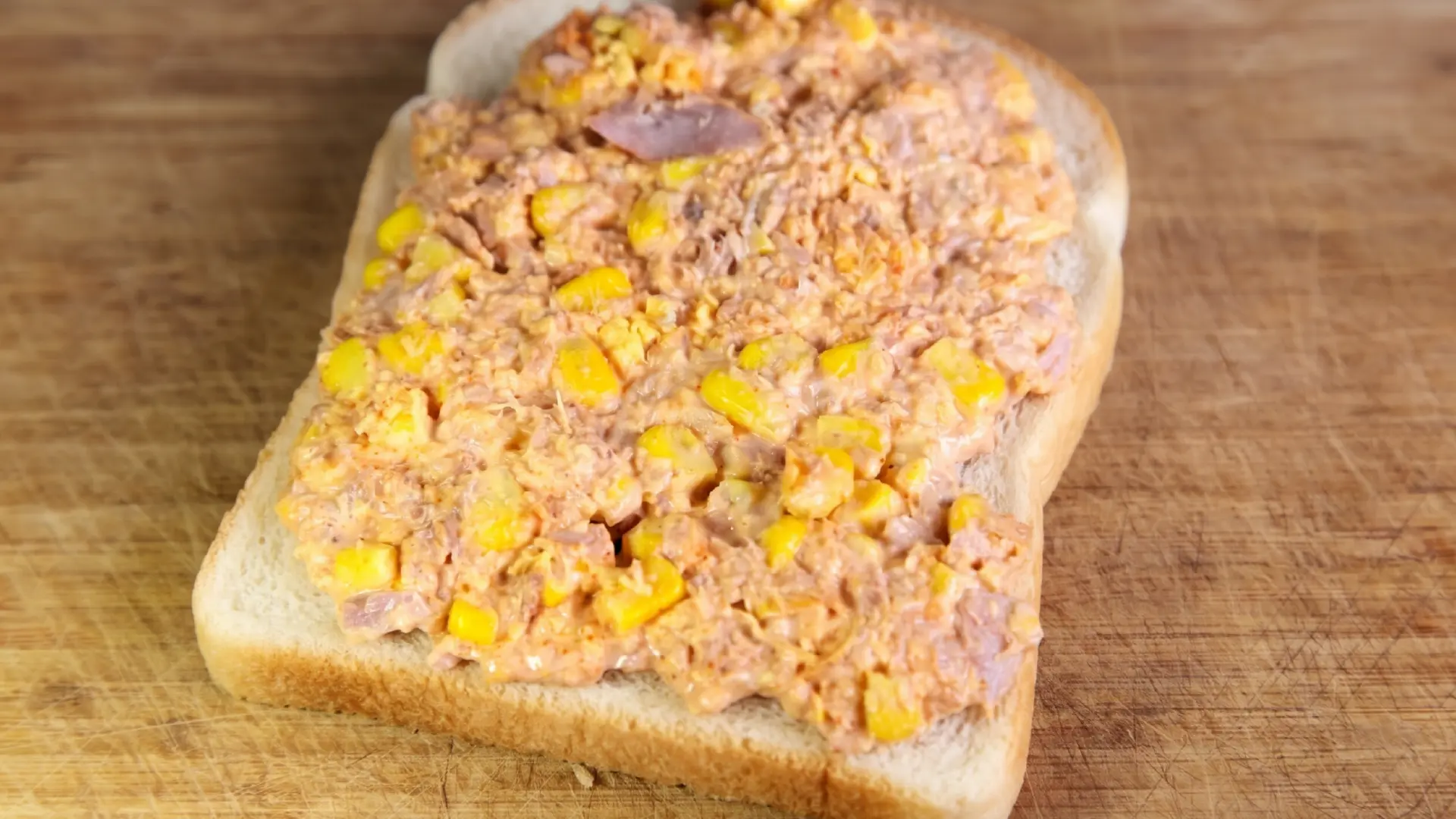 Creamy tuna mayo and sweetcorn filling spread evenly across a single slice of white bread on a wooden board.