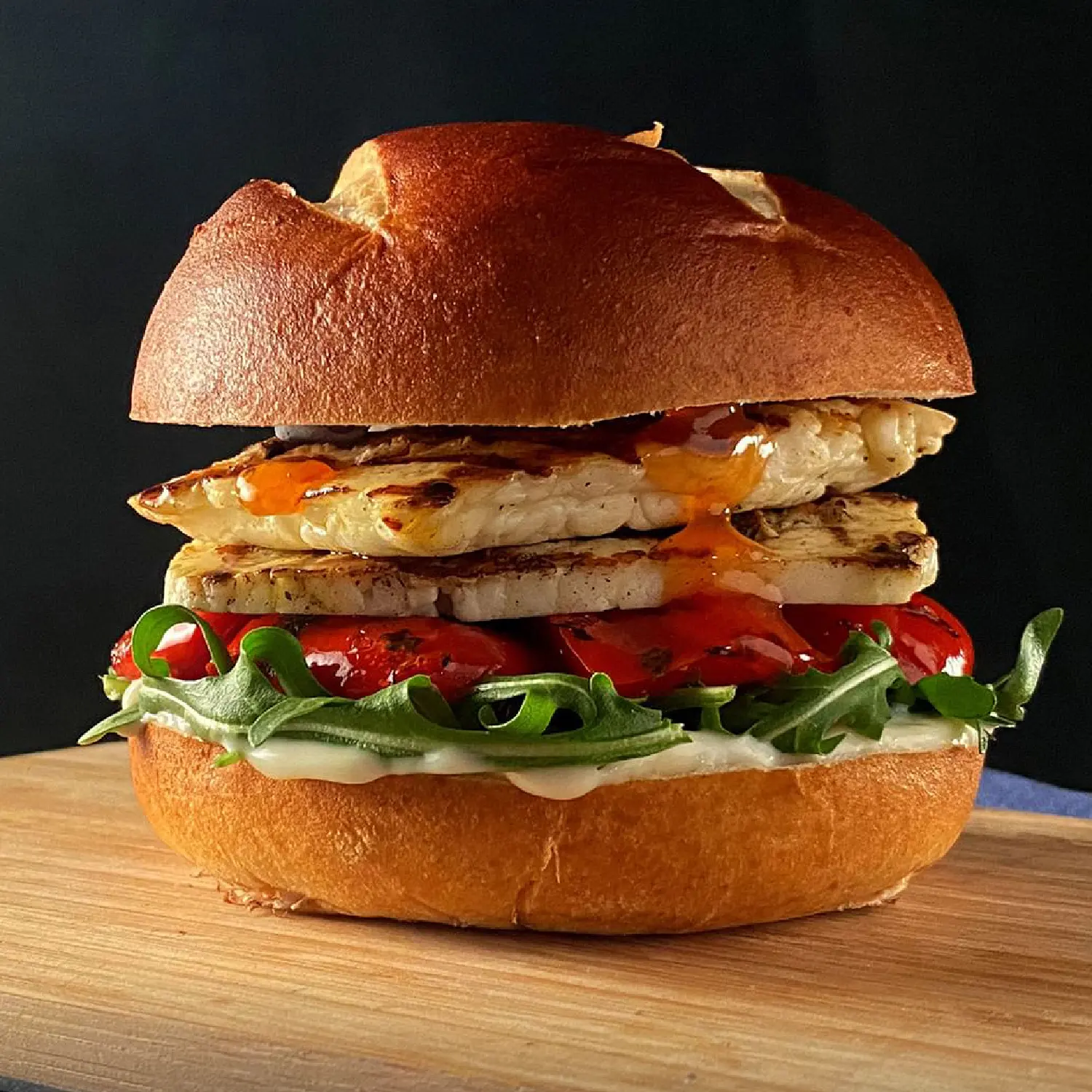 Grilled halloumi burger with sweet chilli, roasted tomatoes and rocket in a brioche bun on a wooden board.