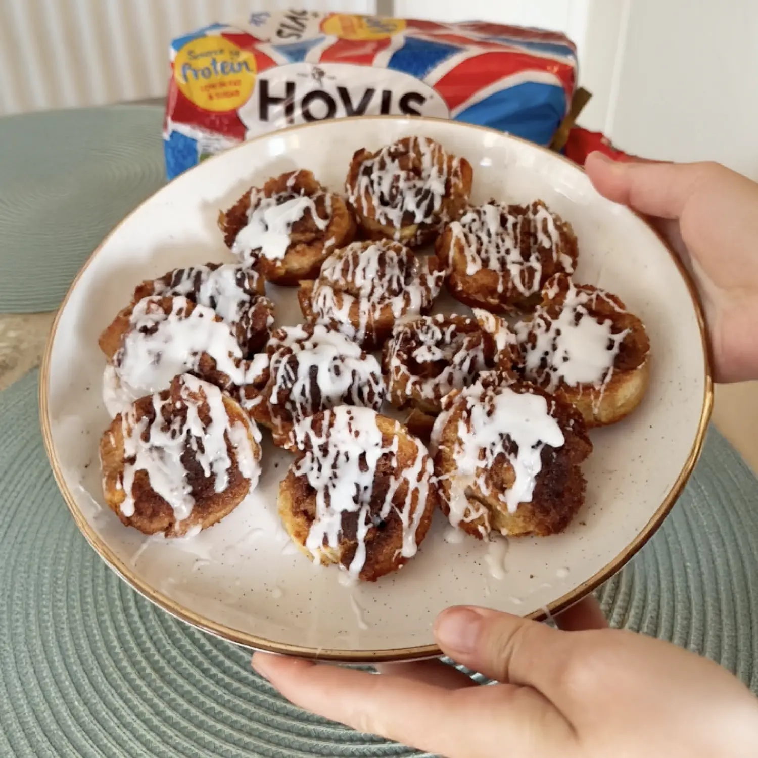 Hands holding a plate of small cinnamon rolls drizzled with white icing, with a Union Jack Hovis bread loaf in the back.