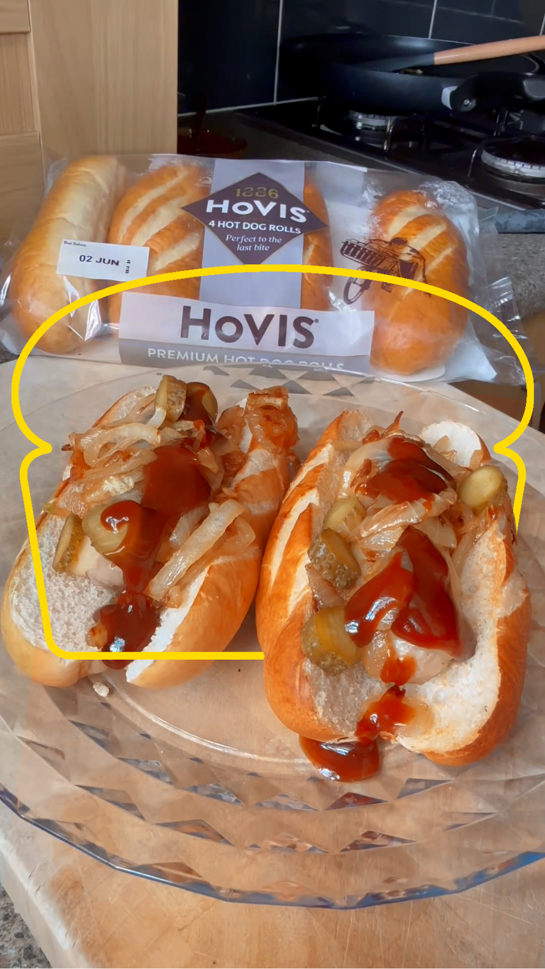 Two hot dogs in soft Hovis hot dog rolls, topped with caramelised onions, sliced pickles, and ketchup, placed on a clear glass plate. In the background, an unopened pack of Hovis hot dog rolls sits on a kitchen counter near a stove.