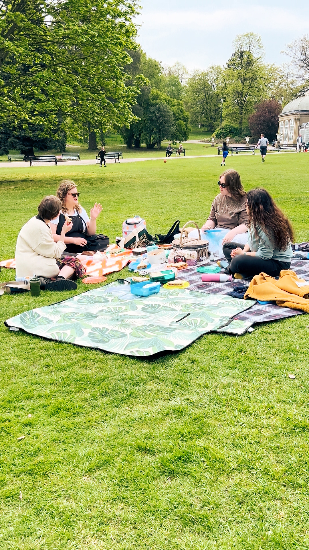 A wide outdoor shot of four people sitting on picnic blankets on a sunny green lawn in a park. They are arranged around a spread of picnic food and drinks. One person is wearing a light-coloured jacket, another a dark top and checked skirt, a third a brown top and sunglasses, and the fourth a dark top. Trees line the background, and other park-goers are visible in the distance, some walking and one running. The overall scene suggests a relaxed social gathering on a pleasant day.