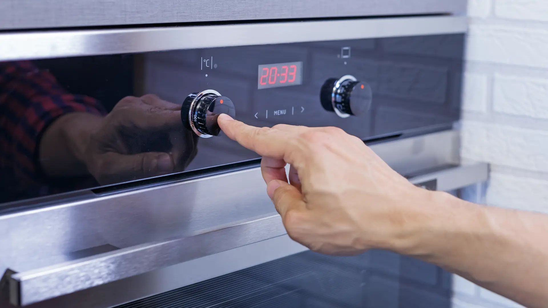 A hand pressing the control knob on a modern stainless steel oven with a red digital display showing 20:33.