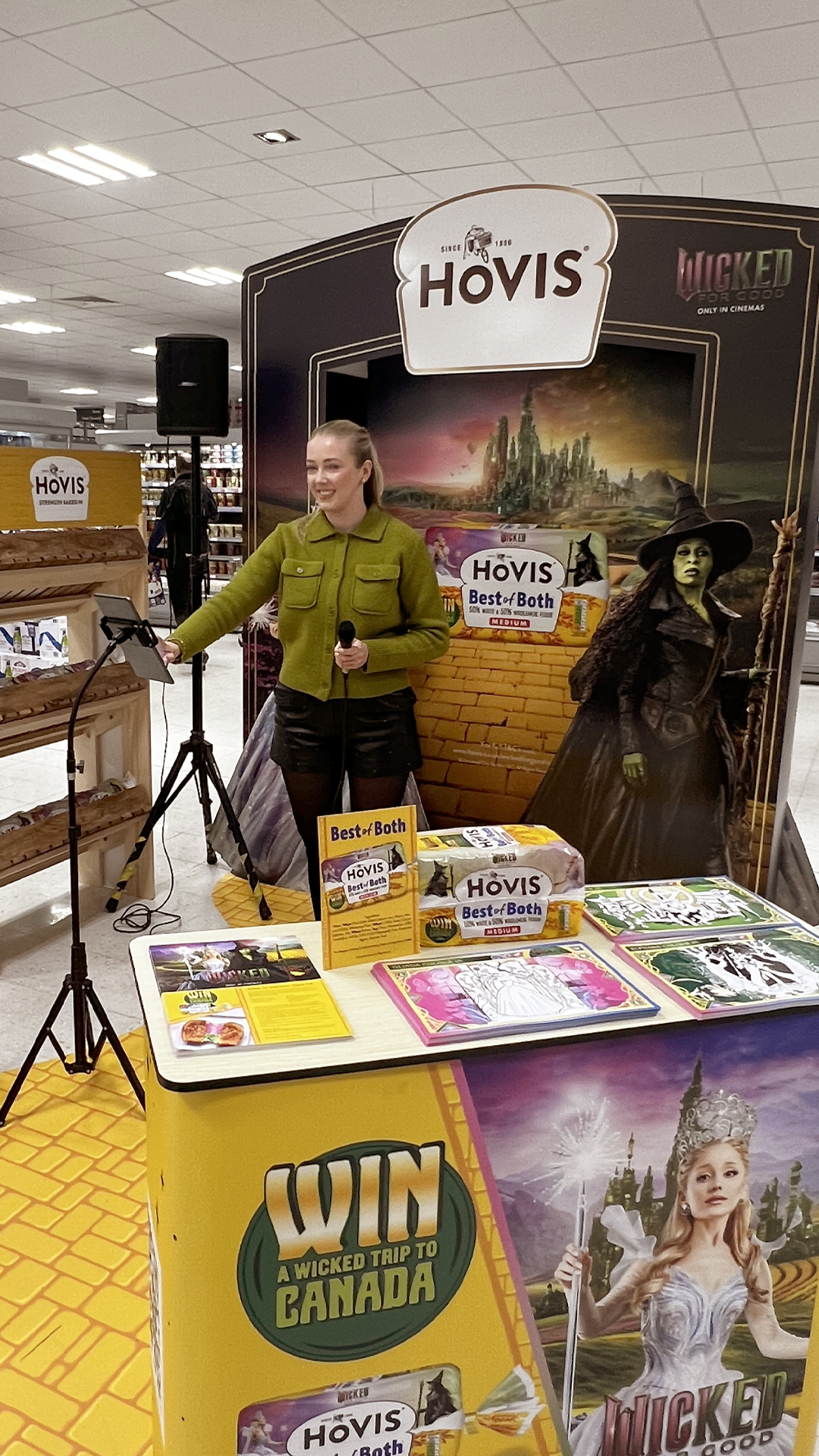 A woman stands at a promotional display for Hovis “Best of Both” bread inside a supermarket. She holds a microphone while smiling and interacting with a tablet on a stand. The setup features a vibrant “Wicked”-themed backdrop, including life-sized character cut-outs and imagery from the film. The counter in front of her is decorated with colourful activity sheets, branded materials, and loaves of Hovis bread, along with a large sign advertising a contest to “Win a Wicked Trip to Canada.”