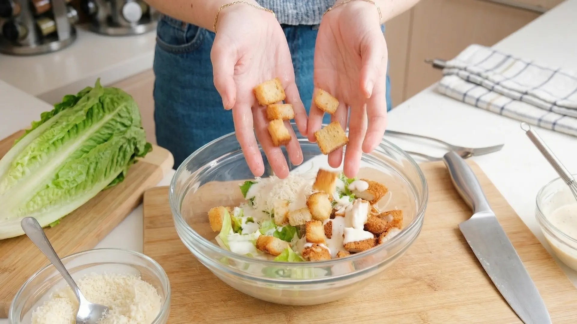 A person drops golden-brown croutons from their hands into a glass bowl of Caesar salad on a kitchen counter.