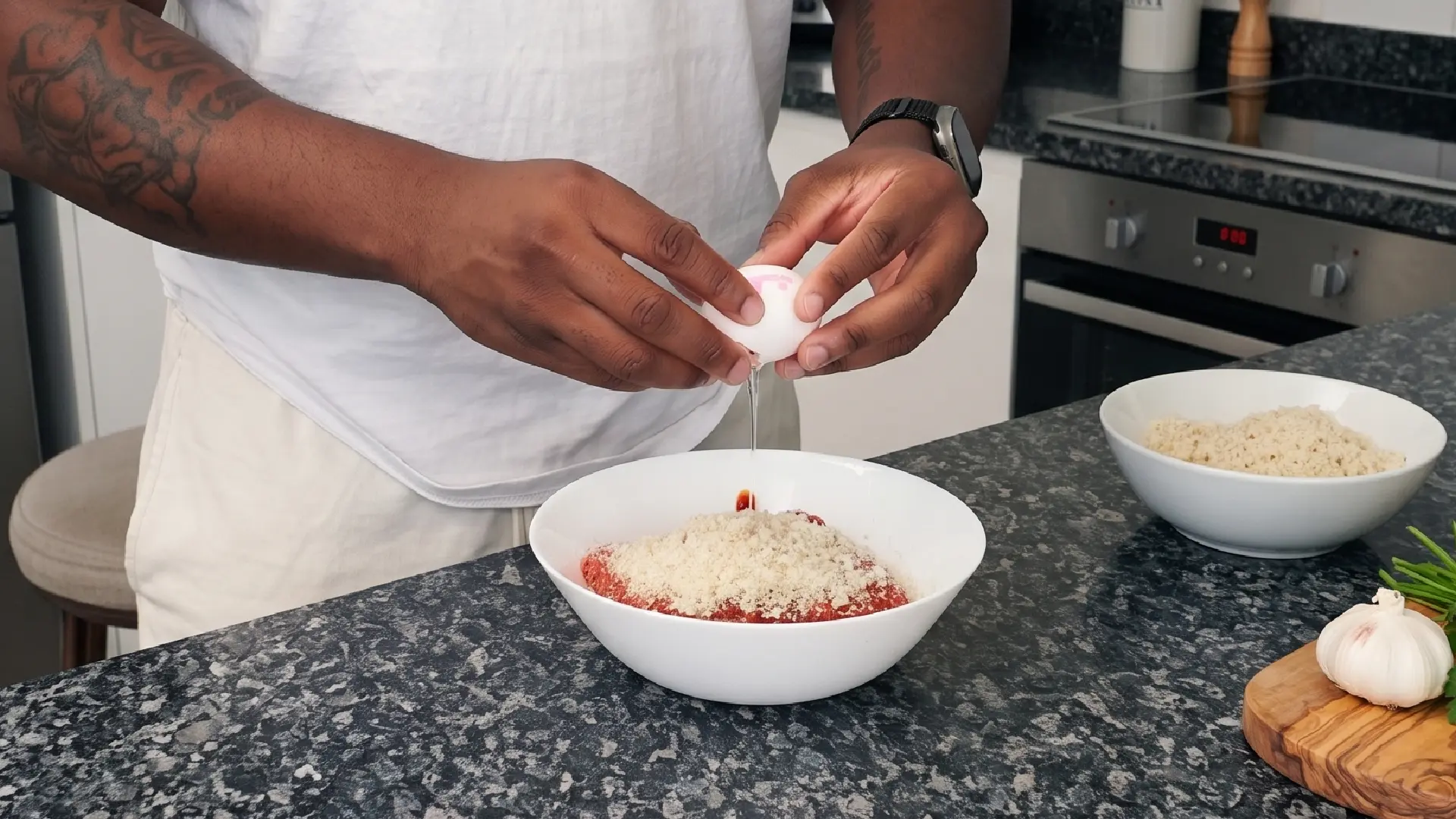 Close up of a person cracking a raw egg into a white bowl containing ground meat and breadcrumbs on a kitchen counter.