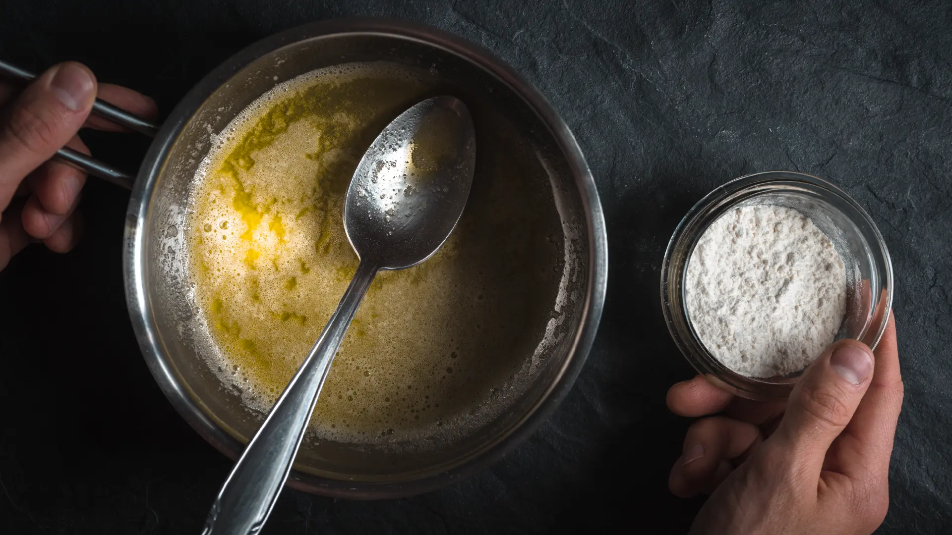 A top-down view of hands holding a bowl of flour next to a saucepan of melted butter, preparing to make a roux.
