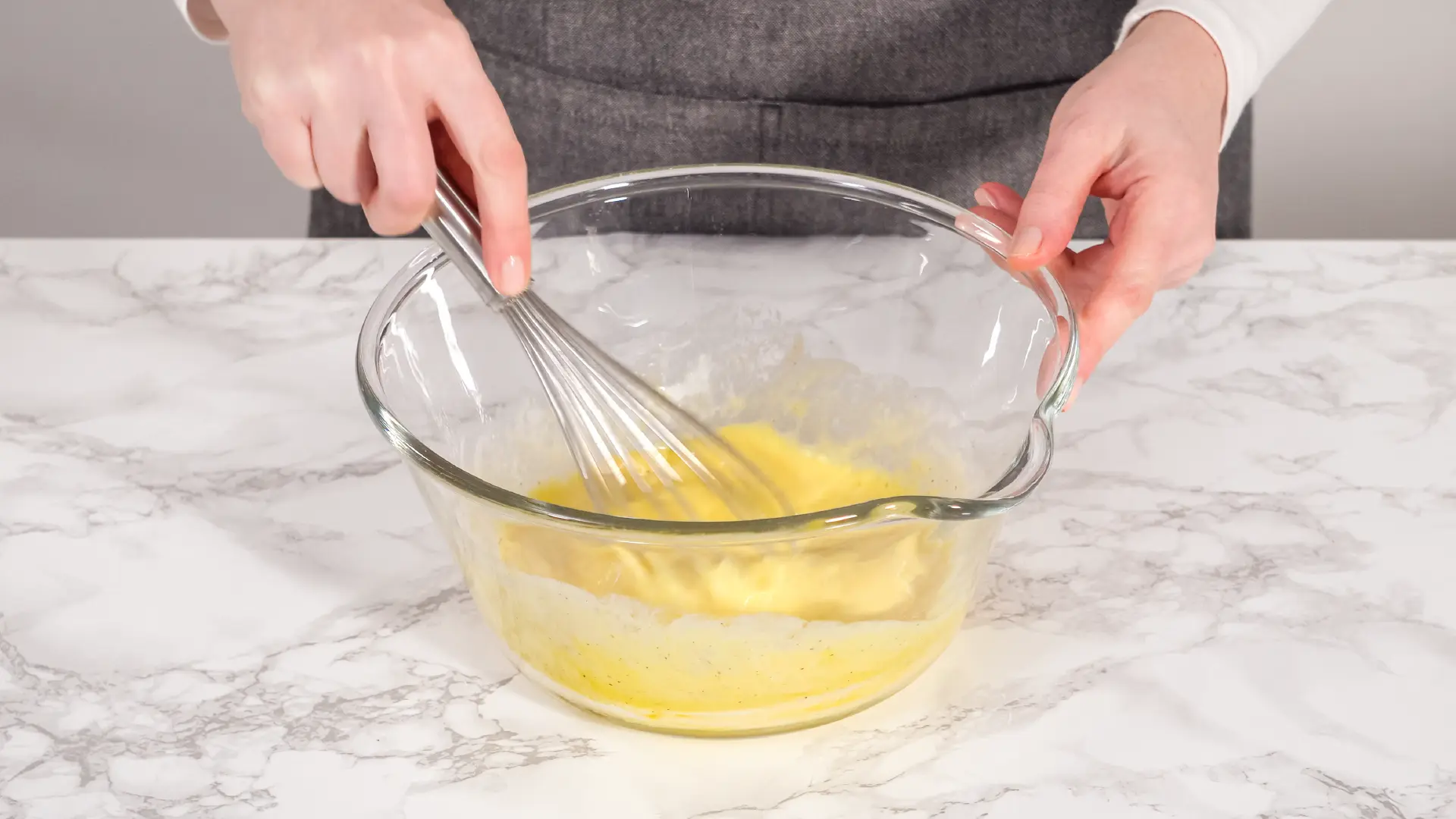 A person uses a metal whisk to mix a yellow batter in a large glass bowl on a white marble countertop.