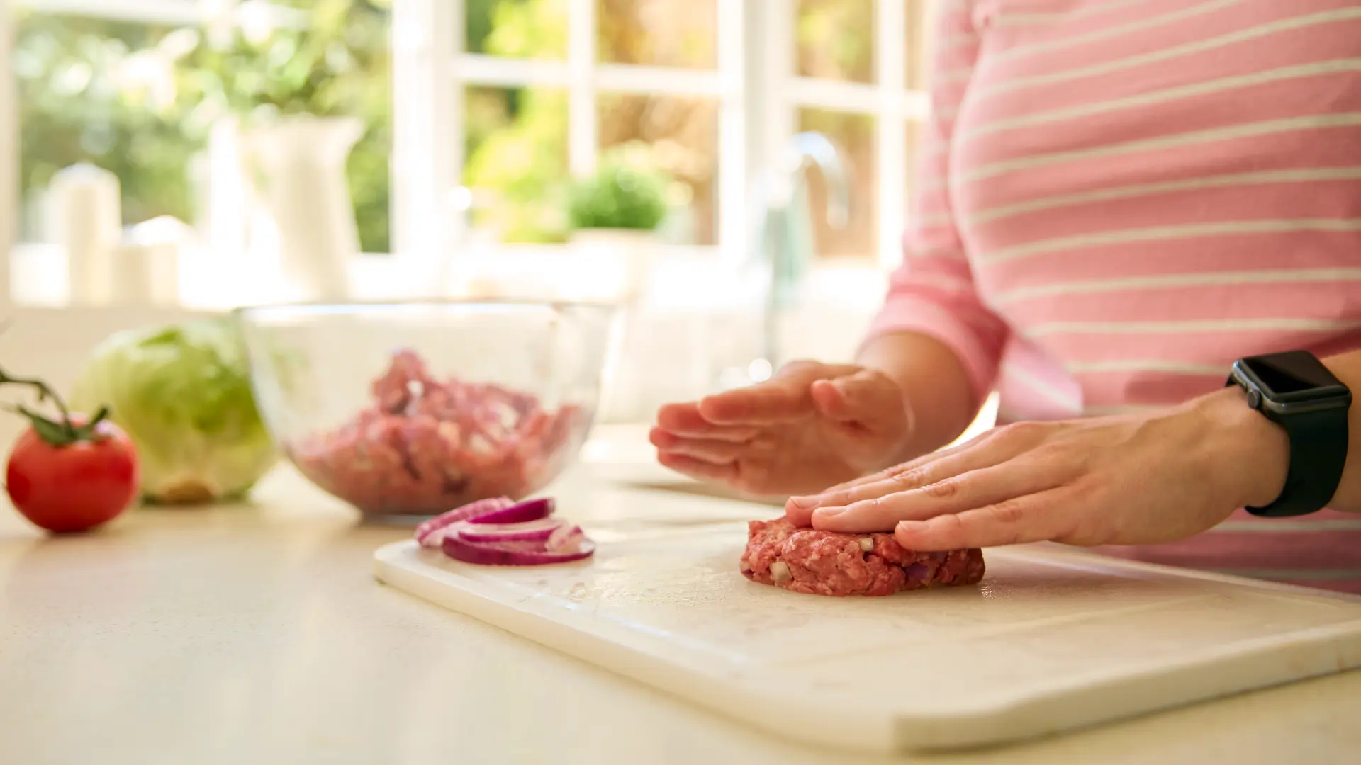 Close-up of hands in a pink striped shirt shaping a raw beef patty on a white board with onions and lettuce nearby.