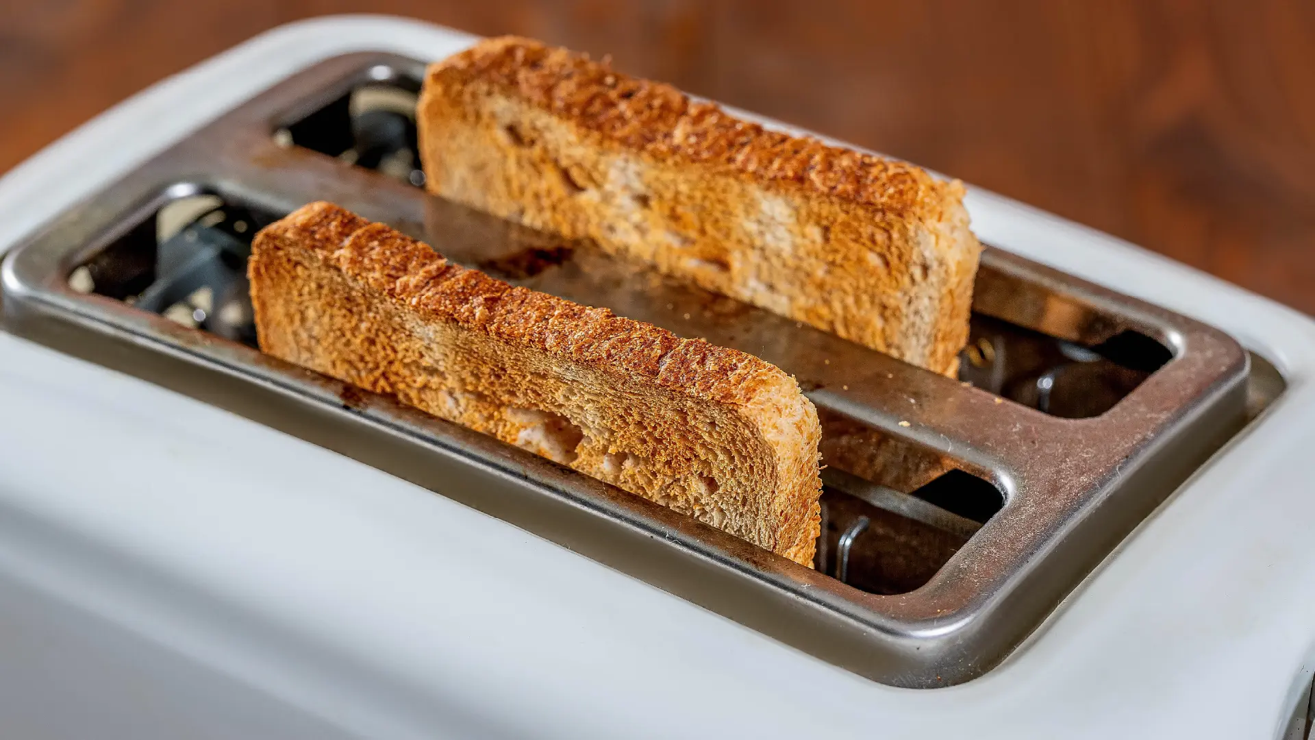 Two slices of golden-brown wholemeal toast popping up from a white two slot toaster with a chrome top.