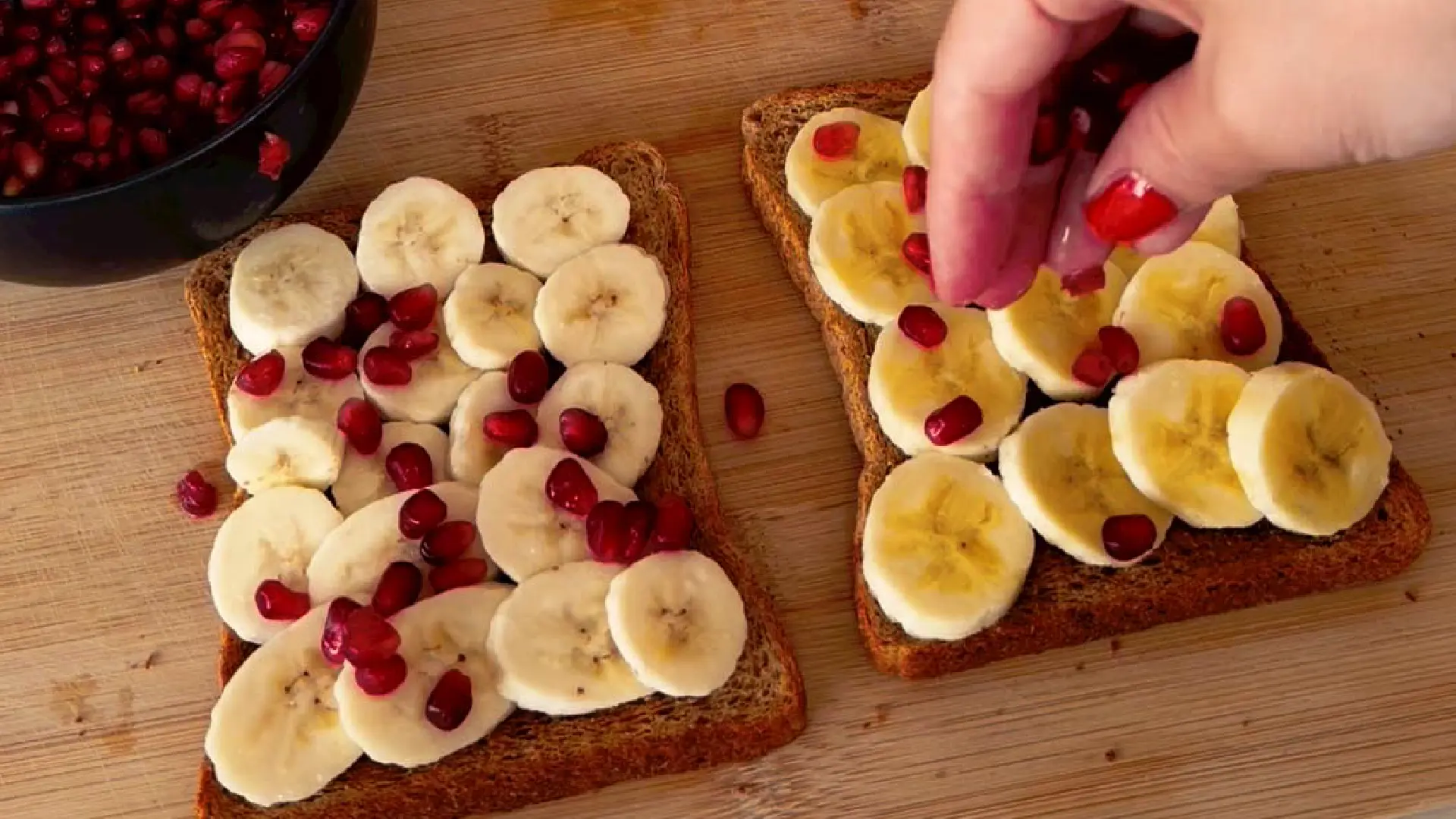 A hand with red nails sprinkling fresh pomegranate seeds over banana topped wholemeal toast on a wooden board.
