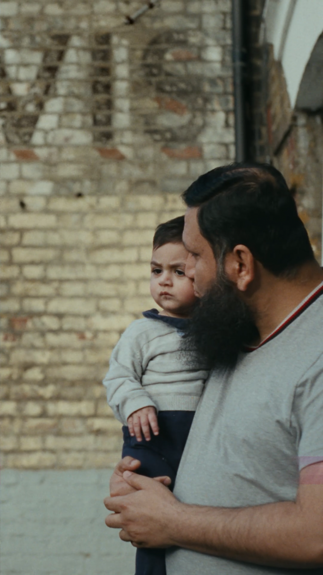 A man with dark hair and a thick black beard holds a toddler in his arms while standing outdoors. A Hovis Ghost Sign in the background.