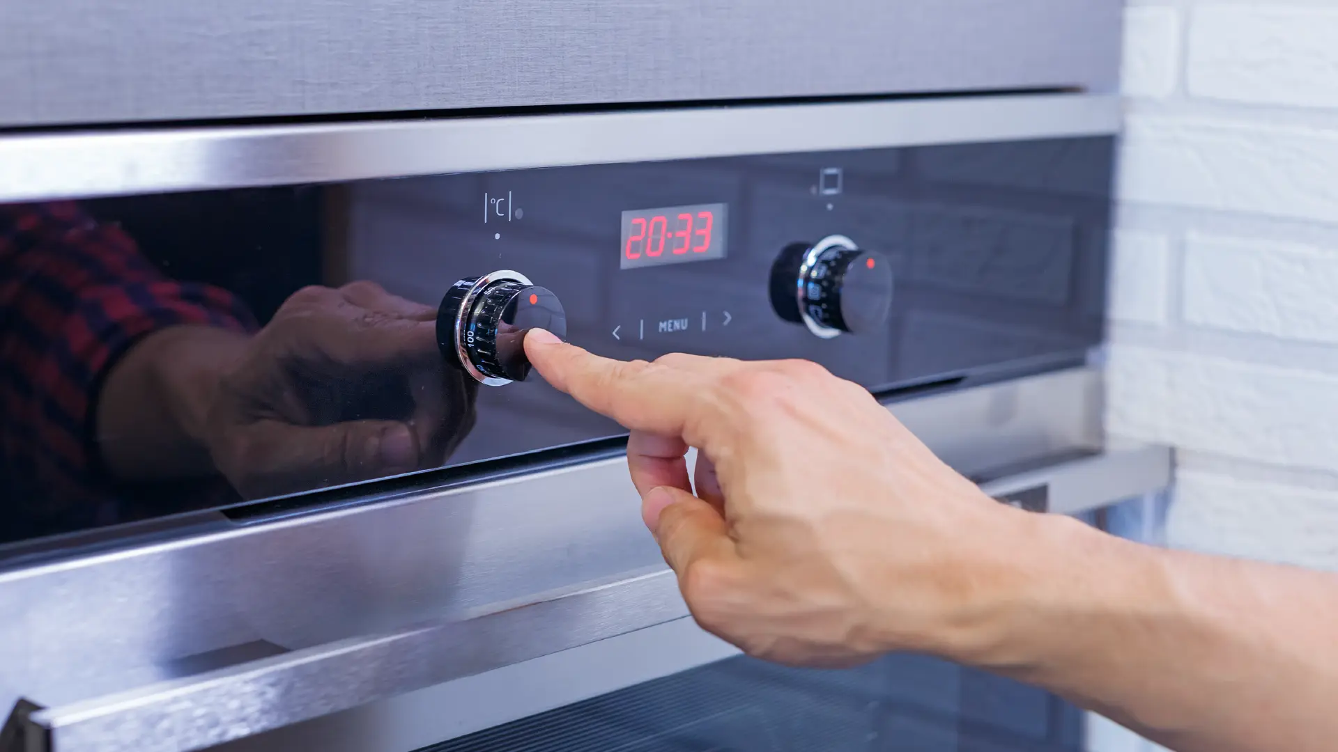 A person’s hand adjusts the temperature dial on a modern stainless steel oven with a digital clock display.